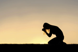 A silhouette of a young Christian woman is bowing her head in prayer, and desperation outside during sunset.